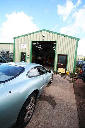 A pale blue Jaguar XK8 parked in front of the workshop building